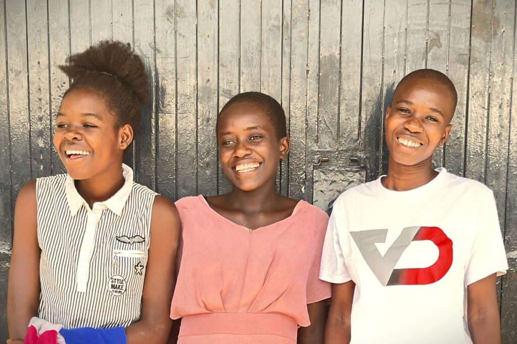 Three young Kenyan women in casual clothes, standing together smiling on a sunny day